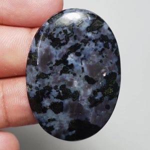 May include: Oval-shaped gemstone with a polished surface. The stone features a mix of dark blue, black, and green patterns. The gemstone is held in a hand, showcasing its size and shape. This stone could be used for jewelry.