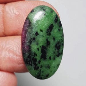 May include: Oval-shaped gemstone with a polished surface. The stone features a vibrant green base color, speckled with black inclusions and a streak of deep red. The gemstone is held between fingers.