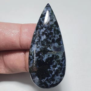 May include: A teardrop-shaped gemstone with a polished surface. The stone features a speckled pattern of dark blue, black, and hints of green. The gemstone is held by a person's fingers against a white background.