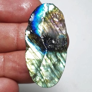 May include: An oval-shaped labradorite gemstone with a mix of colors. The stone has a blue, green, and black iridescence. The surface has a textured appearance with visible lines and patterns.