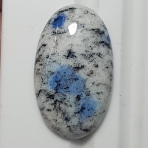 Könnte beinhalten: Ovaler K2-Stein-Cabochon. Der Stein hat eine weiße Basis mit schwarzen und blauen Einschlüssen. Die blauen Einschlüsse konzentrieren sich in der Mitte und oben und erzeugen ein einzigartiges Muster. Der Stein ist poliert und glatt.