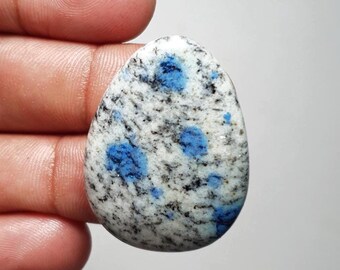 May include: A polished, teardrop-shaped K2 stone. The stone features a white base with speckled black and gray inclusions. Bright blue spots are scattered across the surface, creating a striking contrast. This gemstone is ideal for jewelry making.