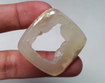 May include: A translucent, square-shaped gemstone with a hollow center. The stone has a light beige color with some areas of white and brown. The edges are smooth and rounded, and the overall appearance is polished.