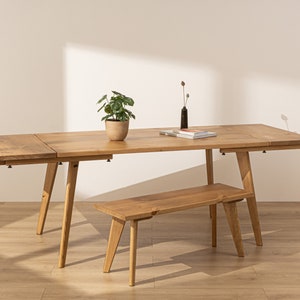 May include: A light wood dining table with a leaf extension and a matching bench. The table has a natural wood finish and is supported by four tapered legs. The bench has a similar finish and is also supported by four tapered legs.