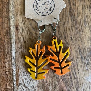 May include: A pair of leaf-shaped earrings with a gradient of orange and yellow hues. The earrings are made of wood and have a dark brown vein design. The earrings are attached to a bronze-colored clasp. The earrings are on a brown tag with the text "Courtney Inspirations.com".