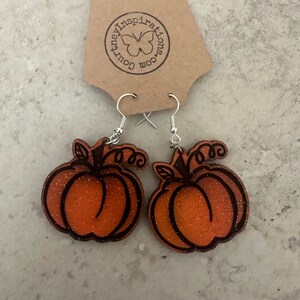 May include: A pair of orange glitter pumpkin earrings with black outlines and silver hooks. The earrings are attached to a brown tag with the text "courtneyinspirations.com" and a butterfly logo.