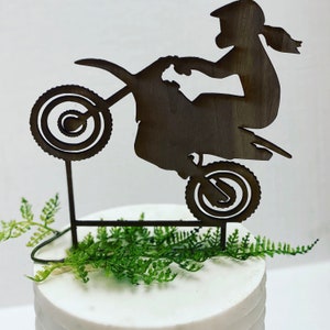 May include: A wooden cake topper featuring a silhouette of a person riding a dirt bike, with the front wheel raised. The topper is brown and has green fern accents. The cake is white.