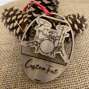 May include: Wooden oval ornament featuring a drum set design with the words "Custom Text" engraved below. The ornament has a red ribbon for hanging. The background includes two pine cones.