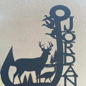May include: Black metal decorative piece featuring a deer, turkey, trees, and flying birds within a hook shape. The name "JORDAN" is vertically displayed. The piece is likely a cake topper or decorative item.
