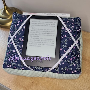 Peut inclure: Un coussin pour liseuse avec un motif floral bleu marine, tenant une liseuse Kindle. Des bandes élastiques blanches maintiennent l'appareil. Le coussin a une bordure en velours côtelé vert clair. Du texte est visible.
