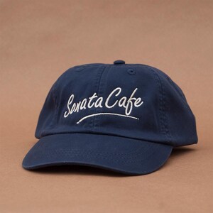 May include: A navy blue baseball cap with white embroidered text that reads "Sonata Cafe".