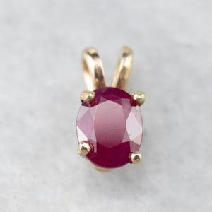 May include: A small, oval-shaped ruby pendant set in a gold setting. The ruby is a deep red color and has a high polish.