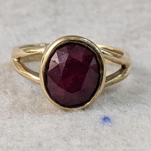 May include: A gold ring with a deep red oval gemstone set in a bezel setting. The ring has a simple, elegant design.