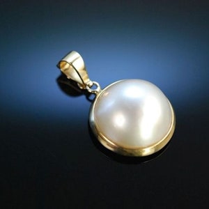 Puede incluir: Un colgante de oro con una perla grande, redonda y blanca. La perla está engastada en un bisel de oro y el colgante tiene un diseño simple y elegante.