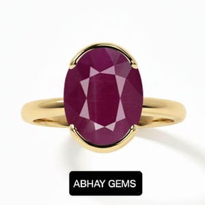 May include: A gold ring featuring a large, oval-cut, deep magenta-red gemstone held in a four-prong setting. The band is smooth and polished gold. Below the ring, a black rectangular label displays the white text ABHAY GEMS in capital letters.
