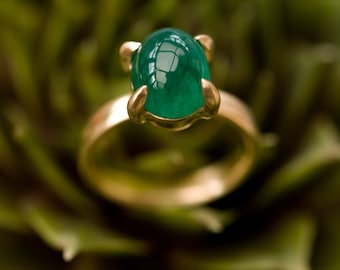 May include: A gold ring featuring a large, oval-cut, vibrant green gemstone. The gemstone is held in place by four gold prongs. The ring band is a simple, polished gold. The background is a blurred, green, leafy texture.