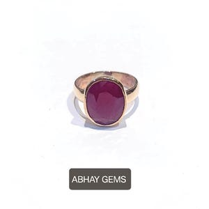 May include: A gold ring featuring a large, oval-cut ruby gemstone. The ruby is a deep, rich red color, set in a polished gold band. The ring is displayed on a white background, with the text "ABHAY GEMS" at the bottom.