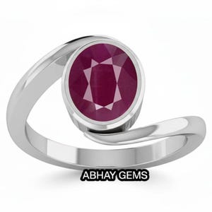 Könnte beinhalten: Ein silberfarbener Bypass-Ring mit einem zentralen, oval geschliffenen, tiefroten Rubin. Die Ringschiene hat eine glatte, polierte Oberfläche, die sich um den Stein windet. Der Text ABHAY GEMS ist in Schwarz unter dem Ring abgedruckt.