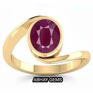 Könnte beinhalten: Ein goldener Bypass-Ring mit einem zentralen, oval geschliffenen, tiefroten Rubin in einer Zargenfassung. Die glatte, polierte Goldschiene schwingt sich um den Stein. Der Text ABHAY GEMS ist unten im Bild in Schwarz aufgedruckt.