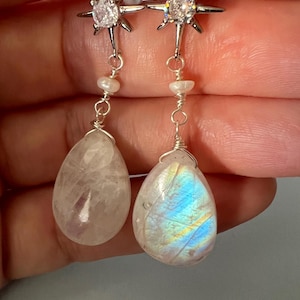 May include: A pair of silver starburst earrings with a clear crystal in the center. Each earring has a white pearl and a teardrop-shaped moonstone pendant.