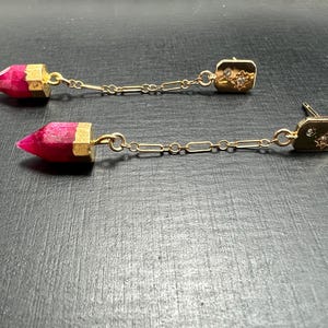 May include: A pair of dangle earrings. Each earring features a gold-colored rectangular stud with a small starburst design and a chain that connects to a faceted, pink gemstone pendant. The earrings are displayed on a dark surface.