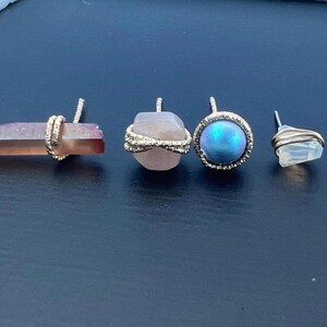 May include: Four gemstone earrings. One is a long, pinkish-orange crystal wrapped in gold wire. Another is a rounded, light pink stone with gold wire. A third is a blue-green iridescent stone in a gold setting. The last is a clear, irregular stone with gold wire.