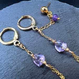 Könnte beinhalten: Goldfarbene Ohrringe mit einem hängenden Design. Jeder Ohrring hat einen kleinen Reifen, eine Kette und einen herzförmigen lila Edelstein. Ein Ohrring hat einen Stecker mit einer kleinen lila Perle.