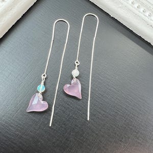 May include: A pair of silver threader earrings. Each earring features a faceted, light pink heart-shaped charm, a small blue-green bead, and a small white bead. The earrings are displayed on a black surface.