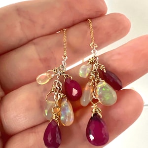 May include: Dangling earrings featuring a cluster of gemstones. The earrings have a gold chain and wire, with a mix of teardrop-shaped ruby and opal stones. The opals have a milky, iridescent quality, while the rubies are a deep red.