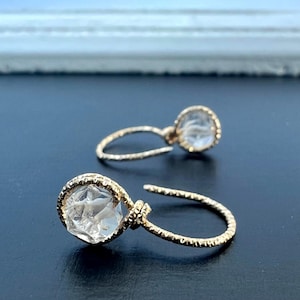 Op de afbeelding: Een paar goudkleurige oorringen met een heldere, facetgeslepen steen in het midden. De oorbellen hebben een textuurontwerp op de ring en een gedetailleerde zetting rond de steen.