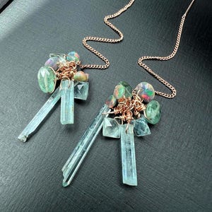 May include: A pair of dangle earrings featuring clusters of colorful gemstones and aquamarine crystals. The earrings have a delicate chain and wire design, with a mix of green, blue, and iridescent stones.