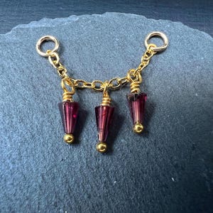May include: A gold-colored chain with three dangling burgundy-colored glass beads. Each bead has a gold-colored cap and a teardrop shape. The chain has two small rings at each end.