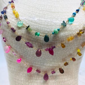 Puede incluir: Collar multicadena con una variedad de piedras preciosas de colores, incluyendo rosa, verde, morado y naranja. El collar está hecho de cadena de oro y presenta piedras preciosas en forma de lágrima.