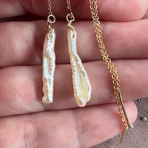 May include: A pair of elegant, long dangle earrings featuring irregular, off-white baroque pearls. The pearls are suspended from delicate gold-colored chains. The earrings are designed to be worn through the ear, creating a stylish and unique look.