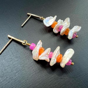 May include: A pair of gold-tone stud earrings. Each earring features a cluster of white, irregular-shaped pearls, interspersed with orange and pink beads. The earrings are set against a dark background.
