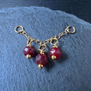 May include: A gold-colored chain with three ruby-colored gemstone pendants. Each pendant is a faceted, round bead with a gold-colored bead at the base. The chain has small loops at each end and in between the pendants.