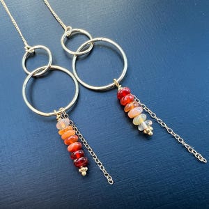 May include: Gold-tone earrings featuring three connected hoops. Each earring has a strand of colorful, graduated beads in shades of red, orange, and yellow, suspended from the bottom hoop. A delicate gold chain hangs below the beads.
