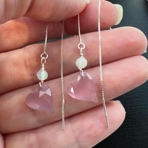 May include: A pair of silver threader earrings featuring faceted light pink heart-shaped stones suspended from thin silver chains. Each earring is accented with a small, round, iridescent white bead positioned directly above the pink heart charm.