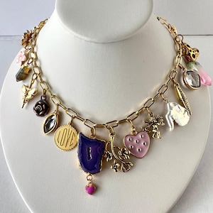 Peut inclure: Un collier à breloques doré avec divers pendentifs, dont une tranche d'agate violette, un cœur rose et un charme avec le mot "LOVE". D'autres breloques incluent un chérubin, une fleur et un cristal.