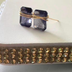 Peut inclure: Boucles d'oreilles à clous avec des pierres rectangulaires violet foncé, enveloppées de fil doré. Les boucles d'oreilles sont posées sur une surface blanche, avec une bordure décorative de strass dorés.