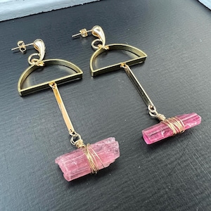 May include: Gold-coloured, geometric drop earrings. Each earring has a half-circle top, a vertical bar, and a pink tourmaline crystal wrapped in gold wire. The earrings have a post-back closure.