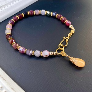 May include: A delicate bracelet made with a variety of colorful gemstone beads, including pink, purple, and blue. The bracelet has a gold clasp and a small, teardrop-shaped pendant.