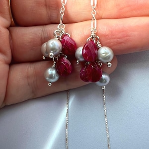 May include: A pair of dangle earrings featuring silver chains with clusters of faceted deep pink ruby-colored teardrop beads and smooth, round, light gray pearls. The components are connected with silver wire wrapping and delicate metal links.
