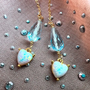 May include: A pair of gold-toned drop earrings. Each earring features a teardrop-shaped, clear blue crystal above a heart-shaped opal pendant. The opal hearts have a shimmering, iridescent quality. The earrings are set against a textured, brown background.