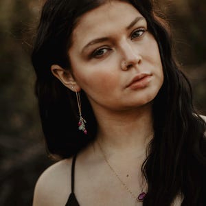 May include: A woman with long dark hair wearing a black top and a gold necklace with a purple stone pendant. She is wearing long gold earrings with pink and green stones.