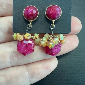 Puede incluir: Un par de pendientes colgantes con una gran piedra de rubí rosa en la parte superior y un racimo de piedras de ópalo más pequeñas que cuelgan debajo. Los pendientes están hechos con alambre de oro y plata.