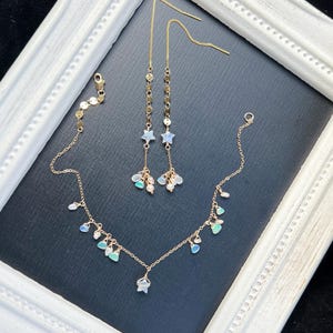 May include: A gold-toned jewelry set featuring a necklace and a pair of earrings. The necklace and earrings are adorned with small, colorful gemstones and star-shaped charms. The jewelry is displayed on a dark surface within a white framed border.