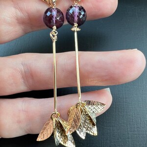May include: Gold-toned dangle earrings featuring faceted purple beads, opal-like accents, and leaf-shaped charms. The earrings have long, slender gold bars connecting the elements, creating an elegant and nature-inspired design.