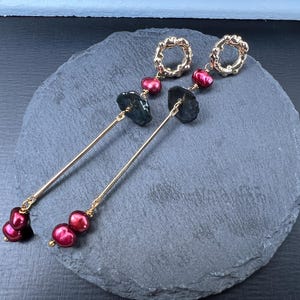 May include: A pair of gold-toned earrings with a textured circular top, a dark gray stone, and a cluster of three maroon-colored pearls. The earrings are displayed on a dark gray, round stone surface.