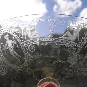 May include: Close-up of a clear glass with etched classical figures and decorative scrollwork. The rim features a series of figures and a central vase design. The glass has a wide, shallow bowl and a rounded base.